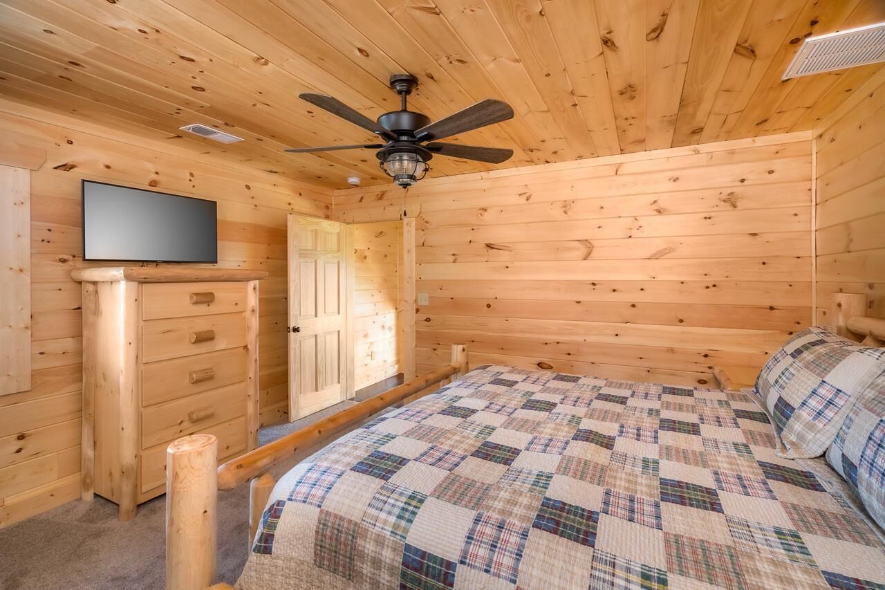 A wooden cabin bedroom with a bed, a dresser, a television, and a ceiling fan.