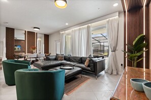 A modern living room with a sectional sofa, green armchairs, a dining area, and a large window with curtains. - Resort Near Disney! Pool! Epic Family Fun! Lazy River! (Clermont)