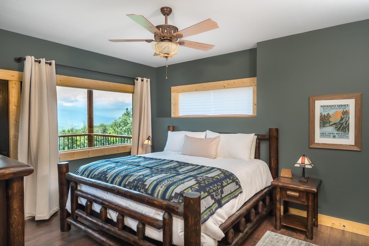 Un dormitorio con cama de madera, ventilador de techo, ventana con cortinas, mesita de noche y un cuadro enmarcado en la pared.