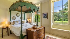 A four-poster bed with a green canopy, a wooden dresser, a window with a view of greenery, and a framed picture on the wall.