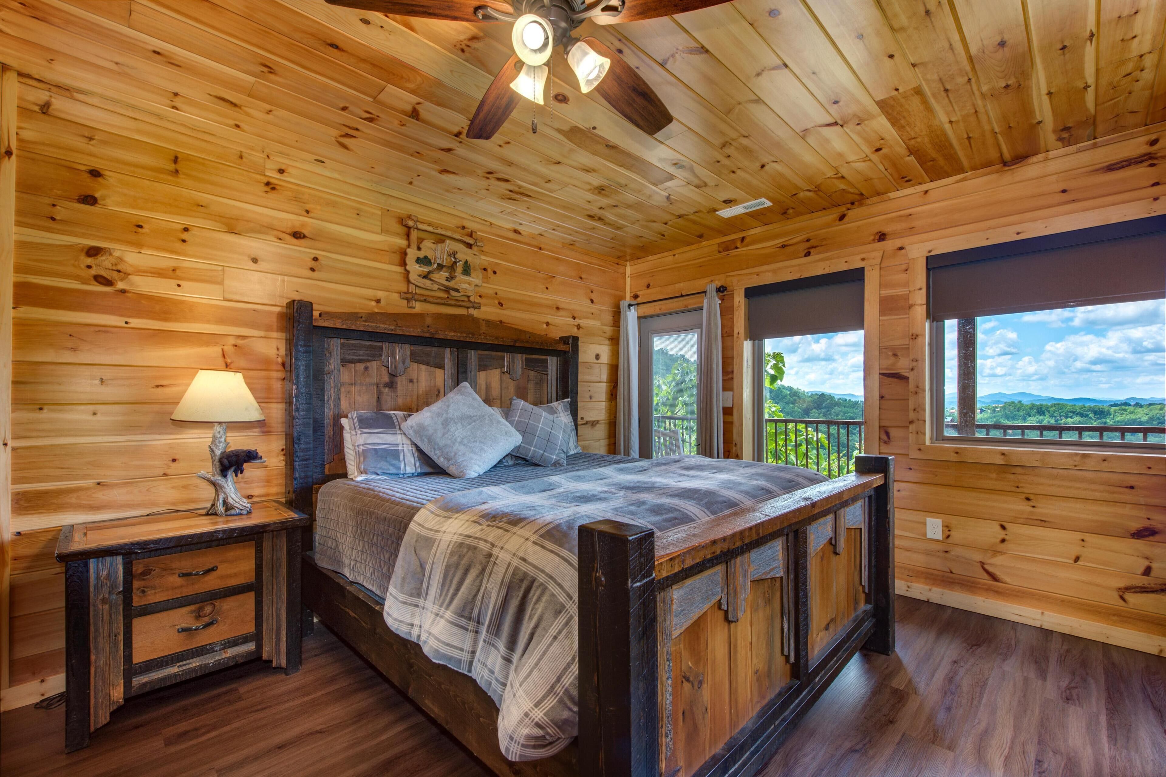 A wooden cabin bedroom with a bed, nightstand, lamp, ceiling fan, and large windows.