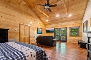Ein Schlafzimmer in einer Blockhütte mit einem Bett, einer Couch, einem Deckenventilator und großen Fenstern.