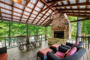 A covered outdoor area with a stone fireplace, wicker furniture, and a view of a forest. - River Lodge (Sevierville)