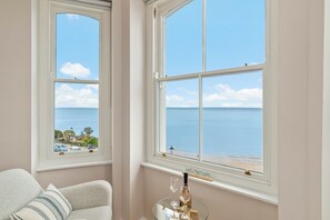 Ein Zimmer mit zwei Fenstern mit Blick auf einen Strand, einem Glastisch mit einer Flasche und Gläsern sowie einem gestreiften Kissen auf einem Sessel.
