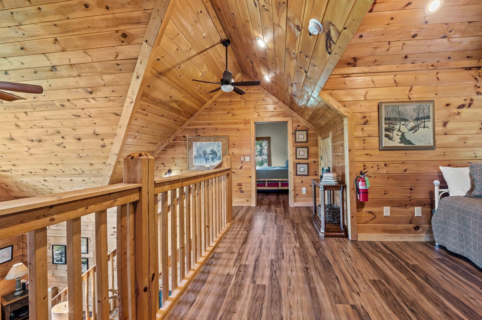 A wooden cabin interior with a wooden ceiling, a ceiling fan, a bed, a fireplace, and a staircase.