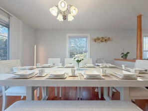 Uma sala de jantar com uma mesa comprida posta para seis pessoas, cadeiras brancas, um candelabro, um relógio na parede e uma janela com vista para árvores.