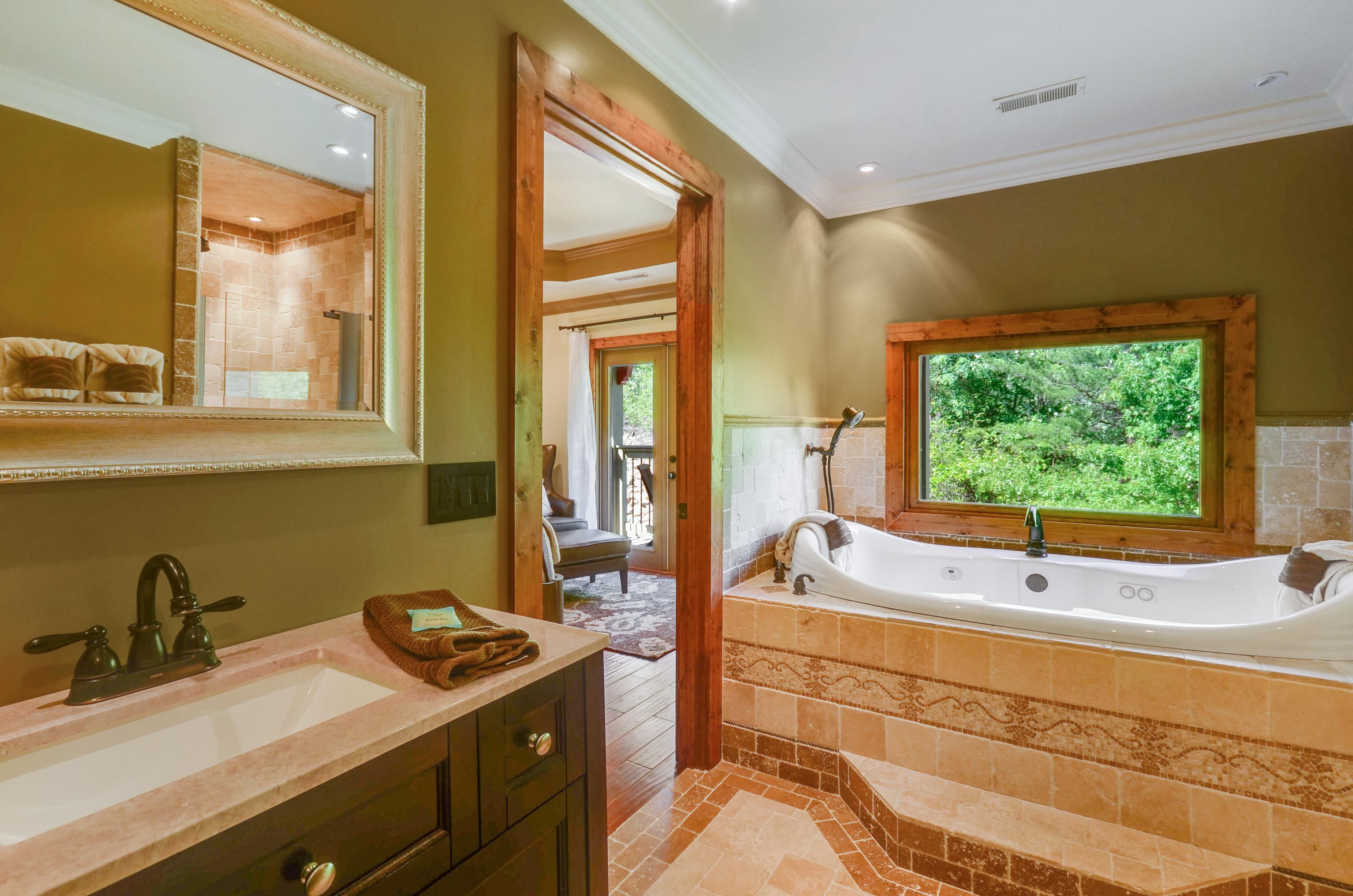 A bathroom with a large mirror, a bathtub, a sink, and a window with a view of greenery.