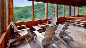 Eine Holzhütte mit großen Fenstern, Sitzgelegenheit im Freien und Blick auf ein bewaldetes Gebiet.