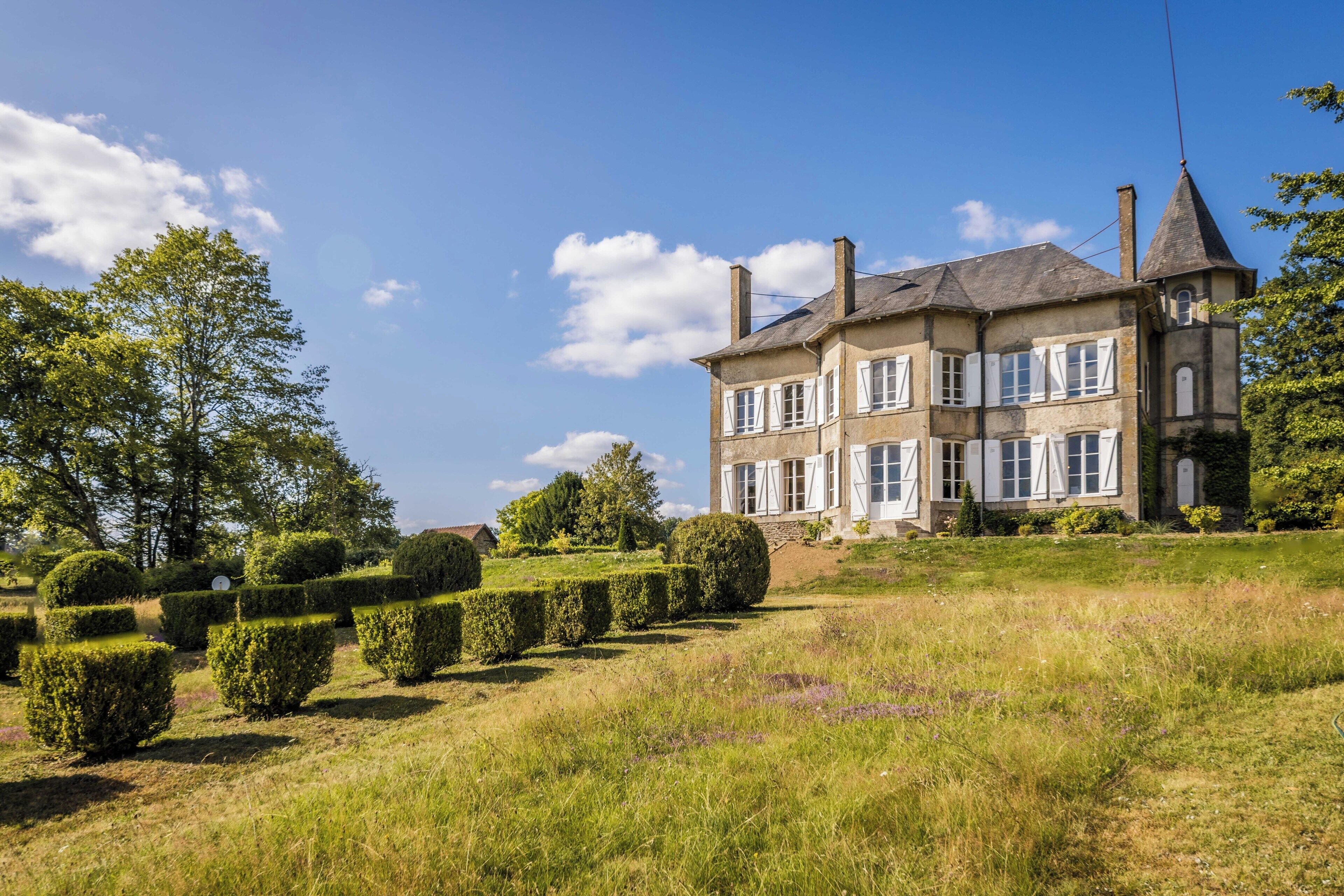 Un grand bâtiment historique, doté de nombreuses fenêtres et d’une tour imposante, entouré de haies bien entretenues et d’une pelouse verdoyante.