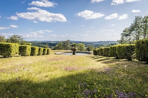 A landscaped garden with a central fountain, neatly trimmed hedges, and a distant view of hills.