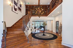 Eine Holztretreppe mit Geländer, eine Chandelüster, ein runder Tisch mit Teppich und ein Bücherregal im Hintergrund.