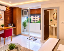 A modern hotel room with a balcony, a desk with a chair, a bed, a bathroom with a sink, and a potted plant.