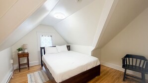Un dormitorio con techo inclinado, una cama, una mesita de noche, un banco y una ventana con cortinas.