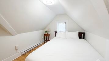 A bedroom with a sloped ceiling, a bed with white bedding, a bedside table with a lamp, and a window with a curtain.