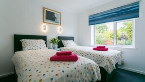 A bedroom with two beds, a window with a view of greenery, a framed picture on the wall, and a bedside table with a vase. - Swn y Mor Bach - sleeps 6 guests  in 3 bedrooms (Criccieth)