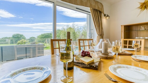 A dining table set with plates, glasses, a bottle, and a candle, overlooking a scenic view. - Swn y Mor Bach - sleeps 6 guests  in 3 bedrooms (Criccieth)