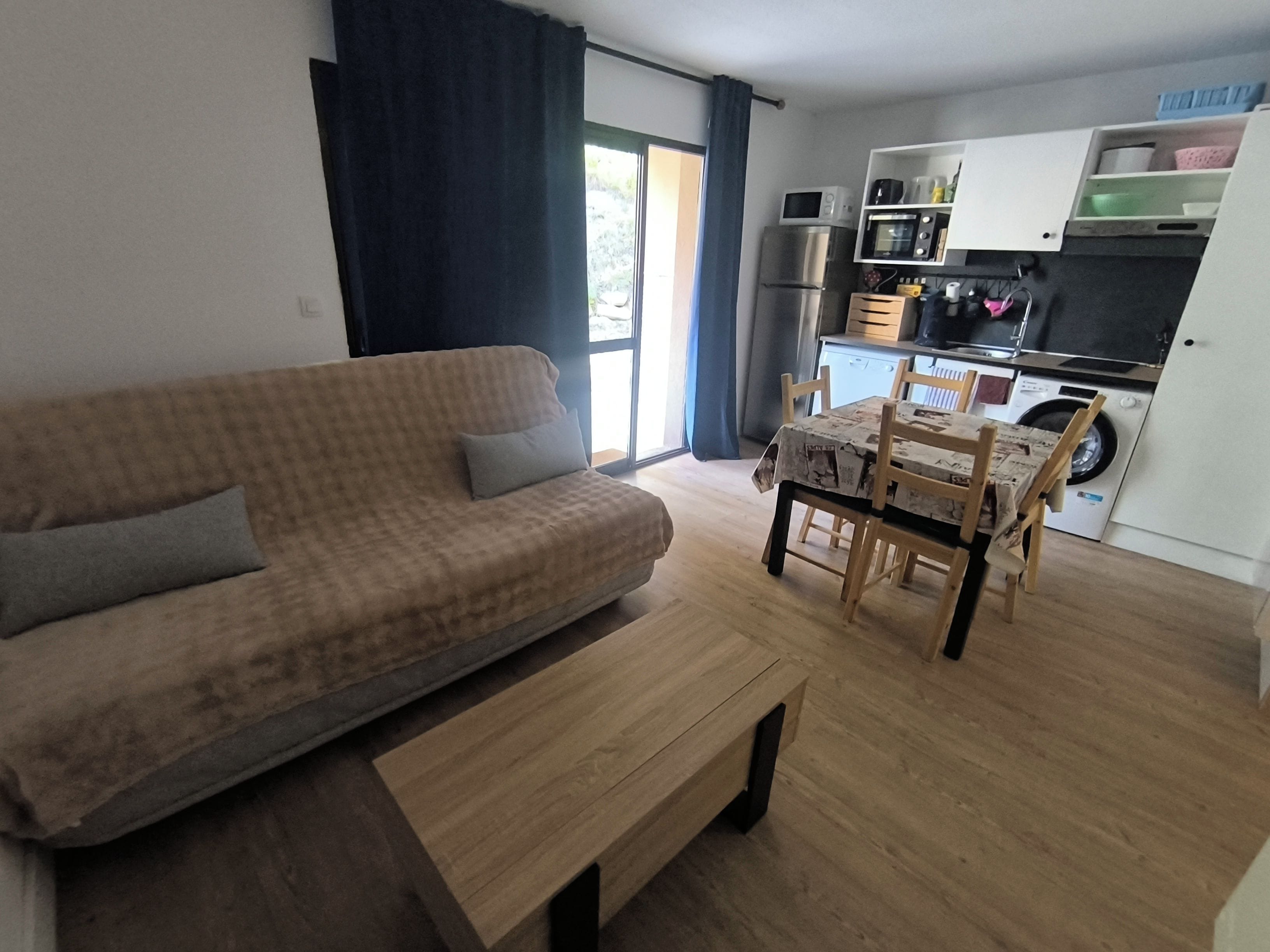 Un salon comprenant un canapé, une table basse et un coin repas avec des chaises. Il y a aussi une cuisine équipée d’appareils électroménagers et d’un lave-linge.