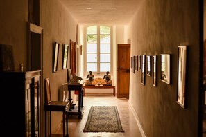 A hallway with framed pictures on the walls, a wooden bench with a fan, and a large window at the end.
