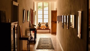 Un couloir avec des tableaux encadrés accrochés aux murs, un banc en bois avec un ventilateur, et une grande fenêtre au fond.