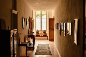 A hallway with framed pictures on the walls, a wooden bench with a fan, and a large window at the end.