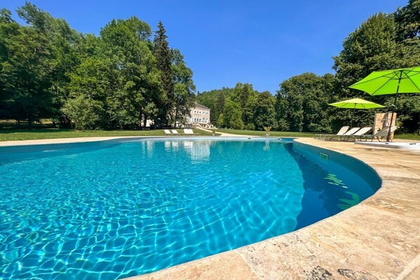 Une piscine aux eaux cristallines, Ă bords incurvĂ©s, entourĂ©e dâarbres et avec un bĂątiment visible au loin.