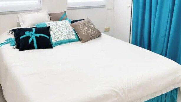 A bed with a white bedspread and teal curtains, a window with blinds, and a wall-mounted air conditioner. - Beautiful 1-bedroom studio with AC in charming Arecibo (Arecibo)