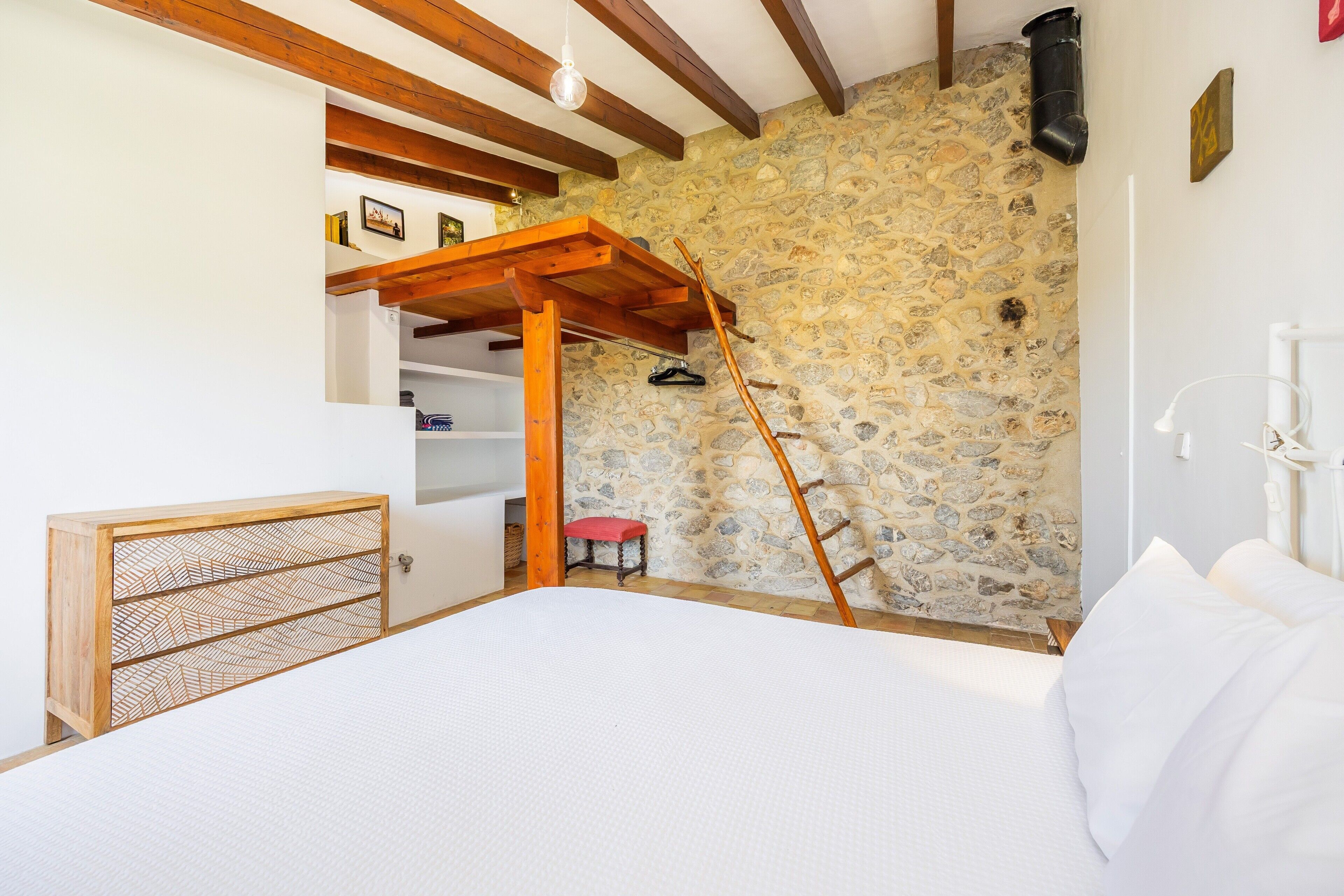 Une chambre avec un mur en pierre, des poutres en bois, un lit, une armoire en bois, un tabouret rouge et une échelle.