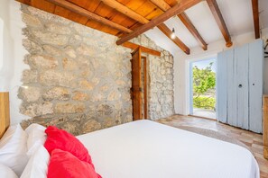 Un dormitorio con pared de piedra, vigas de madera en el techo, una cama con ropa de cama blanca y roja, y vistas al verdor exterior.