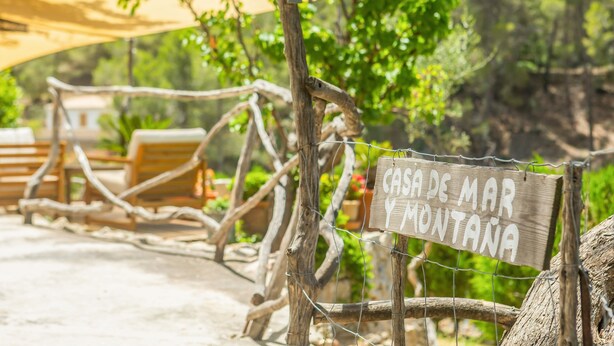 Een rustiek houten bordje met de tekst 'CASA DE MAR MONTANA' bevestigd aan een draadhek, omringd door natuurlijke begroeiing en houten meubels.