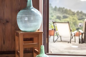 Un jarrón grande y transparente sobre un taburete de madera, cerca de una ventana, con vistas a sillas y follaje al aire libre.