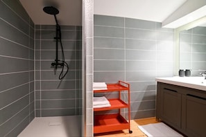 Un baño con paredes de azulejos grises, una ducha, un lavamanos blanco, un espejo y un estante rojo con toallas.