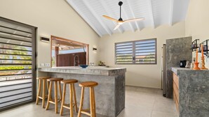 Une cuisine moderne avec un coin bar, des tabourets en bois, un ventilateur de plafond et de grandes fenêtres.