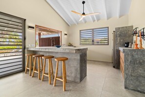 Eine moderne Küche mit Bar, Holzstühlen, Deckenventilator und großen Fenstern.