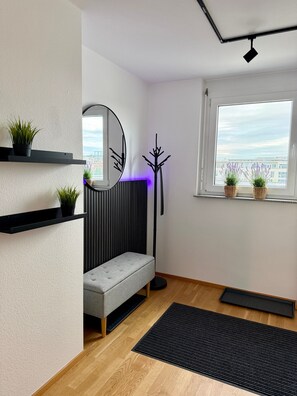 Una habitación moderna con un sofá gris, una lámpara de pie negra, un espejo redondo y dos ventanas con plantas en macetas.