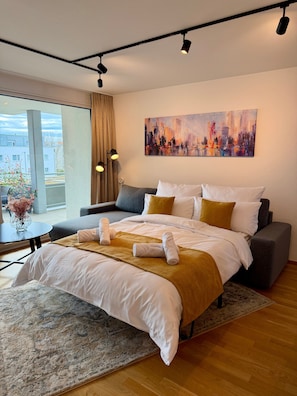 A modern hotel room with a large bed, a sofa, a painting on the wall, and a view of the city through the window.