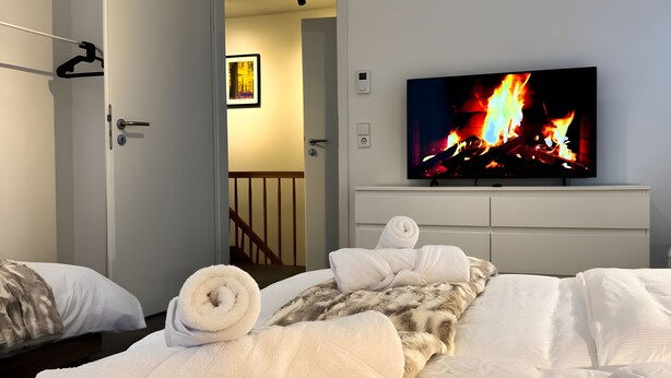Een slaapkamer met een bed, een televisie waarop een haard te zien is, een deur en een gang.