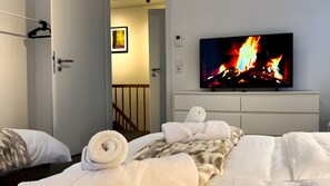 Un dormitorio con cama, televisor mostrando una chimenea, una puerta y un pasillo.