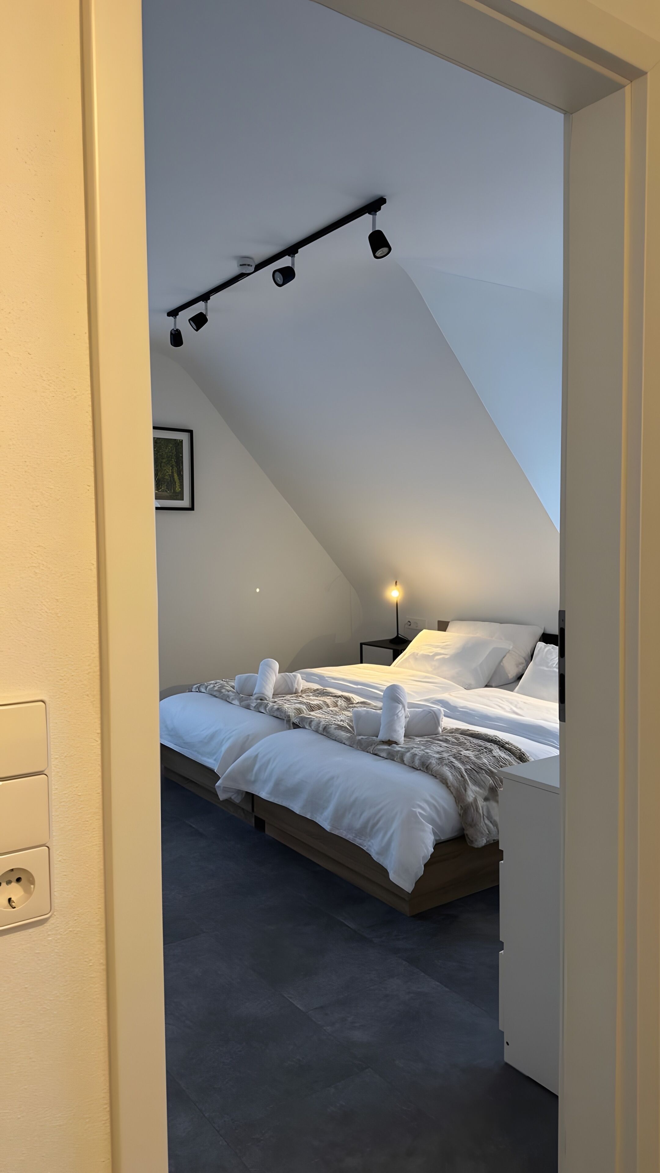 A bedroom with a sloped ceiling, a bed with white linens, a bedside lamp, and a wall-mounted light fixture.