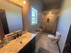 Et badeværelse med vask i marmor, mørke skabe, et stort spejl, et vindue, en bruseniche med stenvæg og et toilet.