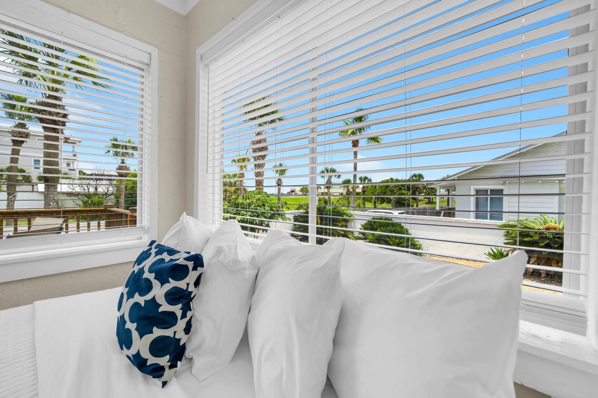 A hotel room with a bed, white blinds, and a view of palm trees and buildings.