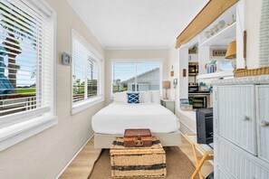 Ein Schlafzimmer mit einem Bett, zwei Fenstern mit Jalousien, einem Nachttisch, einer Lampe, einer Kommode und einem kleinen Tisch mit einem Koffer.