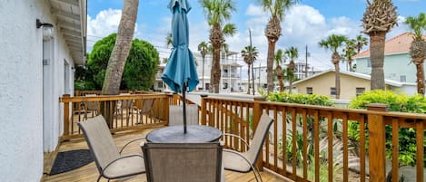 A wooden deck with two chairs and a table, surrounded by a wooden railing, palm trees, and a clear sky.