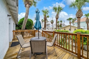 A wooden deck with two chairs and a table, surrounded by a wooden railing, palm trees, and a clear sky. - Linda's Cozy Cottage B (Panama City Beach)