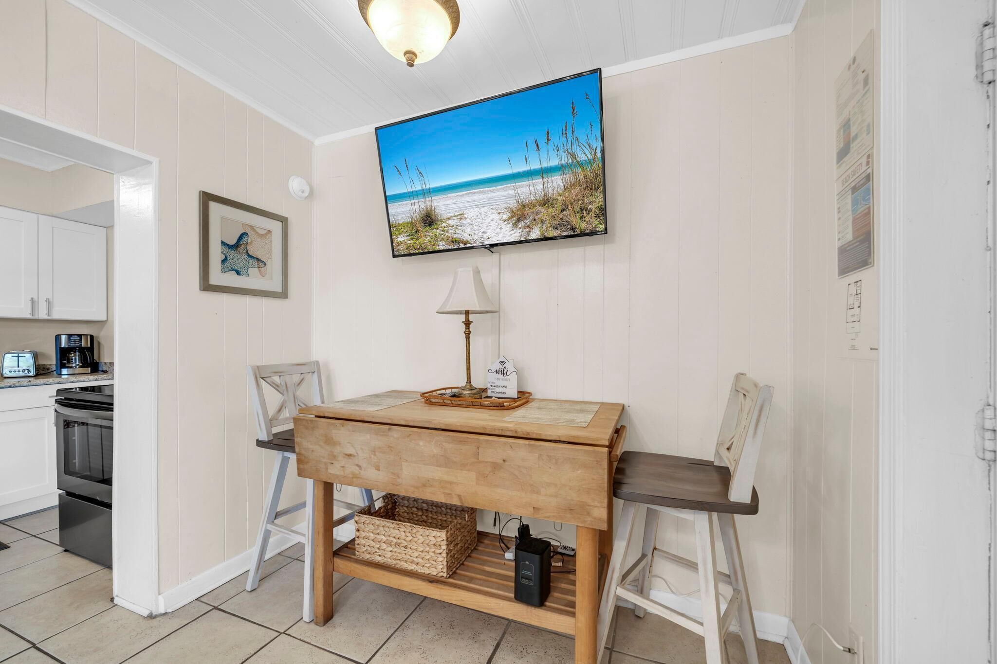 Une pièce comprenant une table en bois, deux chaises, un téléviseur fixé au mur, une lampe et un coin cuisine avec des placards et des appareils électroménagers.