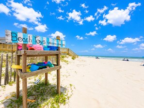 Ein hölzernes Schild mit der Aufschrift 'Beach Toys' und 'Nimm was – lass was' steht an einem Strand mit Holzzaun und einem hölzernen Weg, der zum Strand führt.
