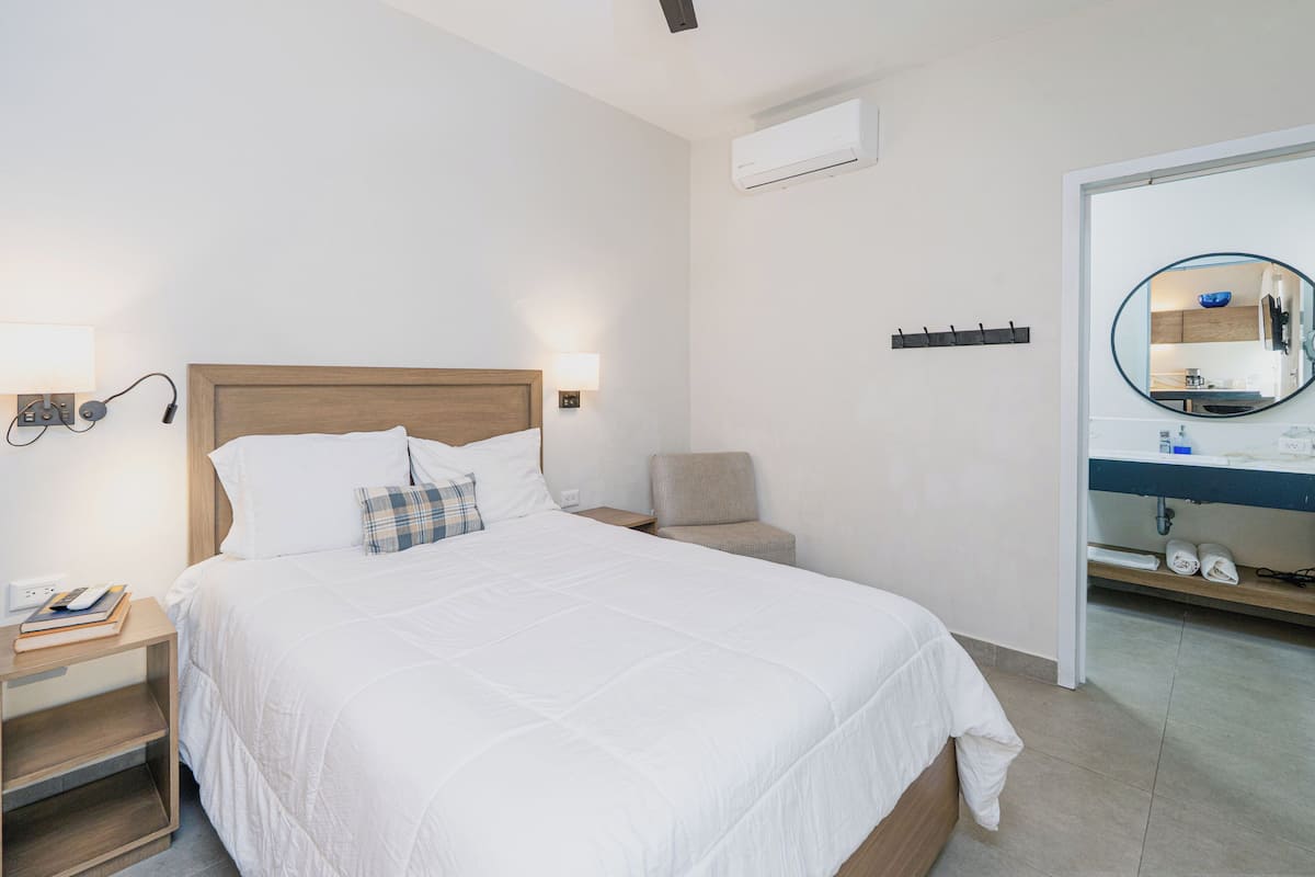 A hotel room with a bed, bedside table, lamp, chair, and a bathroom with a mirror and sink.