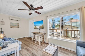 A room with a ceiling fan, a TV, a wooden table with chairs, a sofa, and a large window with blinds. - Linda's Cozy Cottage A (Panama City Beach)