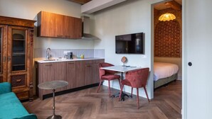 A compact kitchen with wooden cabinets, a dining area with red chairs, a TV mounted on the wall, and a visible bed in the adjacent room. - Studio Urbain (Alkmaar)
