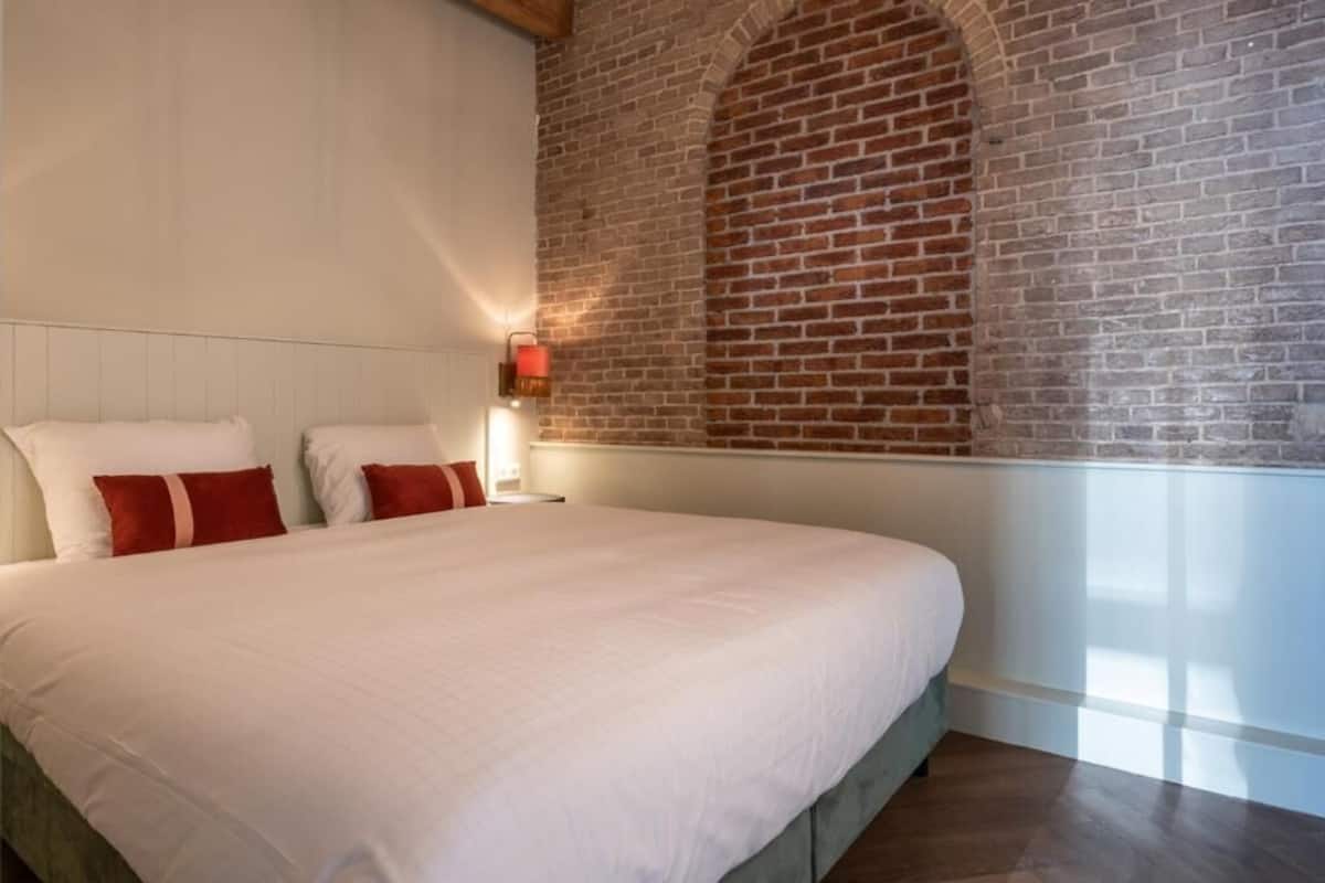 A bedroom with a large bed, white bedding, a brick wall, and a nightstand with a red lamp.
