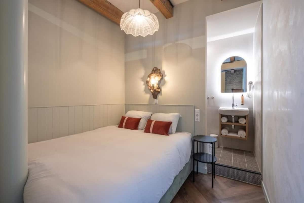 A bedroom with a bed, a bedside table, a wall-mounted light fixture, and a bathroom with a sink and mirror.